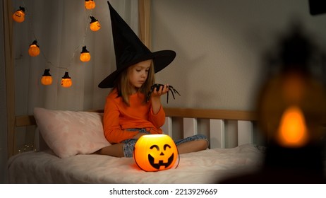 Girl In Witch Black Hat Plays With Spider In Dark With Orange Pumpkin Lantern. Happy Child Celebrats Halloween