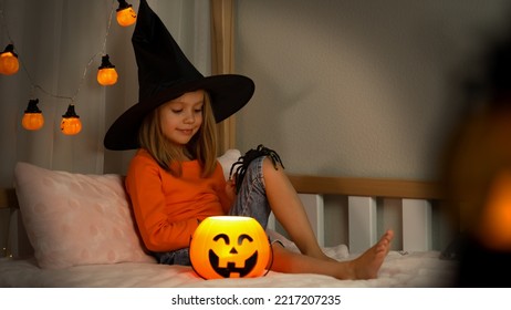 Girl In Witch Black Hat Plays With Spider In Dark With Orange Pumpkin Lantern At Home. Happy Child Celebrats Halloween