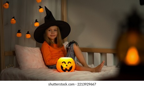 Girl In Witch Black Hat With Spider Looks At Camera In Dark With Orange Pumpkin Lantern At Home. Happy Child Celebrats Halloween