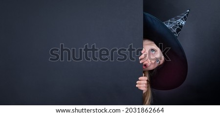 A girl in a witch costume on a black background peeps out from behind the wall. Festive banner with place for your design. halloween