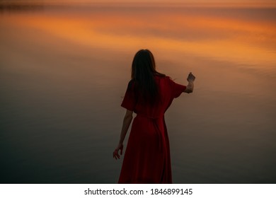 Girl or woman from a back is dancing and praying in a red dress on a background of an orange sunset over the lake in autumn with copy space and text area