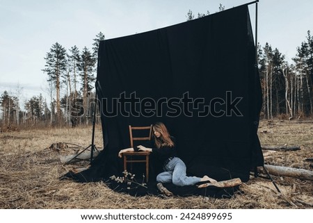 A girl, a woman with bare feet and black clothes sits on the ground on a black background, set among a deforested forest. Wood logs in the background. The wind blows your hair.