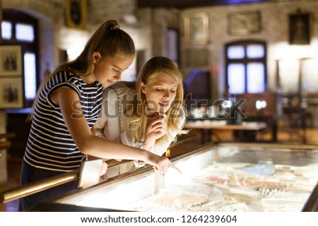 Girl with woman looking with interest at art objects under glass in museum, using guidebook