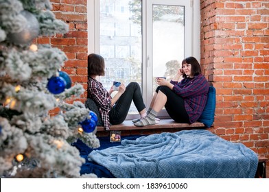 A girl and a woman with mugs sitting on the windowsill in a warm cozy house in winter, the concept of communication, Christmas tree decorated