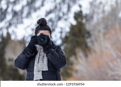 Girl and woman outdoor holding a camera DSLR to her face taking a picture. Outdoor during winter with a mountain and snow background. 