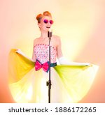 Girl or woman singer singing karaoke into retro vintage microphone. Happy stylish teenager musician in pink glasses smiles, dressed funny dress on colored rainbow background