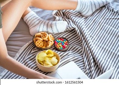 Girl Works At A Computer In A Bed And Eats Fast Food. Unhealthy Food: Chips, Crackers, Candy, Waffles. Junk Food, Concept.