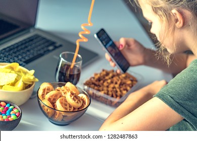 Girl works at a computer and eats fast food. Unhealthy food: chips, crackers, candy, waffles, cola. Junk food, concept.
