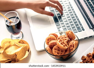 Girl Works At A Computer And Eats Fast Food. Unhealthy Food: Chips, Crackers, Candy, Waffles, Cola. Junk Food, Concept.