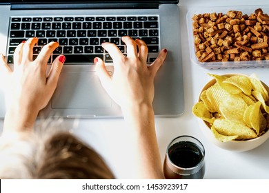 Girl works at a computer and eats fast food. Unhealthy food: chips, crackers, candy, waffles, cola. Junk food, concept.