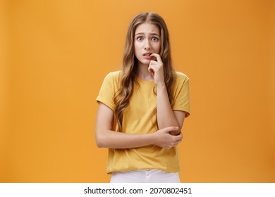 Girl Worried Mom React On New Tattoo. Portrait Of Anxious And Nervous Silly Insecure Young Female With Wavy Natural Hairstyle Biting Finger Panicking Looking Concerned And Troubled Over Orange Wall