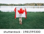 Girl wrapped in large Canadian flag by Muskoka lake in nature. Canada Day celebration outdoor. Kid in large Canadian flag celebrating national Canada Day on 1 of July.