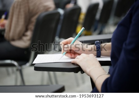 A girl writes a dictation or fills out documents in the audience, sitting on a school chair with a writing stand. Close-up. No face