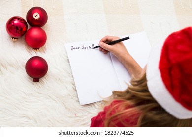 Girl Writing Letter For Santa Claus. Child In Red Santa Hat Lying On Bed And Writing Christmas Letter. Christmas Celebration Concept. Top View Over Shoulder, Coyp Space.