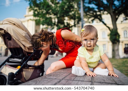 Girl in yellow dress sits on bench while her mother takes something from baby carriage