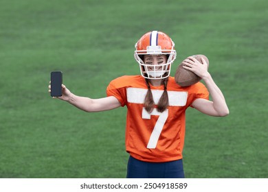 A girl or young woman in a helmet and an American football uniform is holding a ball in one hand and showing a smartphone with the screen facing you in the other. Green grass in the background. - Powered by Shutterstock