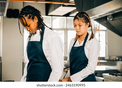 Girlfriend chef helps woman friend tie apron in a restaurant kitchen. Two adults in uniform prepare for food education standing with a painting ready for service. - Powered by Shutterstock