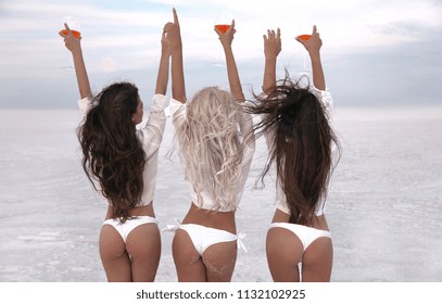 Girlfriend. Happy three female friends in sexy bikini having fun on white beach at sunset. Group of slim ladies cheering with glass of aperol spritz cocktail. Women Enjoying life with drinks.