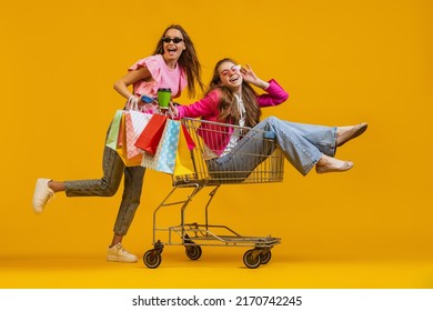 Girlfriends After Shopping. Smiling Young Excited Girls Go Shopping With Shopping Cart Isolated On Bright Yellow Background. Concept Of Sales, Black Friday, Discount, Emotions. Copy Space For Ad, Text