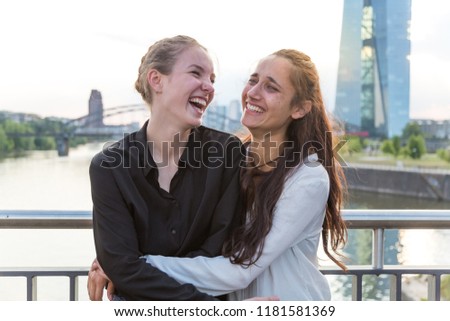 Girlfriends laughing wildly and embracing on city bridge over river with metropolis skyline in the background. Medium shot. 