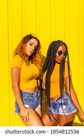 Girlfriends lifestyle, black girl with long braids and blonde Caucasian in yellow shirts and short jeans on a yellow background. Urban session with warm colors in the city