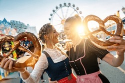 Girlfriends Look To Each Other With Pretzel Or Brezen On A Bavarian Fair Or Oktoberfest Or Duld In National Costume Or Dirndl