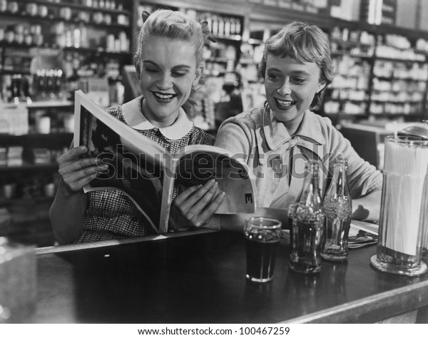 Girlfriends looking\
at magazine at soda\
fountain