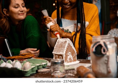 Girlfriends Making Gingerbread house and cookies for Christmas market sale. Women decorating traditional cakes with glaze in late night on kitchen counter