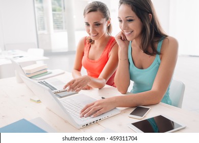 Girlfriends sitting at desk, studying together and smiling, one is pointing at the computer screen