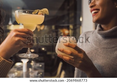 Girlfriends talking together drinking cocktails at restaurant - closeup of young women holding margarita and vodka sour standing at the bar restaurant counter. Focus only hands - unrecognizable faces