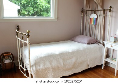 Girl's Bedroom. Iron Frame Canopy Bed With Small Desk Lamp On A Night Stand. A Small Baby Carriage Is In The Room For Pretend Play. Big Open Space Window For Light.