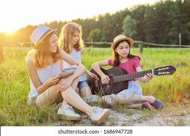 Girls With Classical Guitar On Nature. Children Relaxing On Lawn, Learning To Play The Guitar, Singing Songs, Sunset On Summer Meadow Background