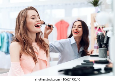 Girls Do Makeup In The Showroom. Two Beautiful Girls Have Fun And Smile. They Are Sitting In A Bright Room Opposite The Mirror.