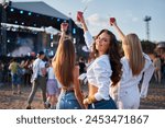 Girls with drinks enjoy live music at sunny beach festival. Group of friends dance, celebrate with cocktails. Summer party vibe on sandy shore. Attendees immerse in concert atmosphere.