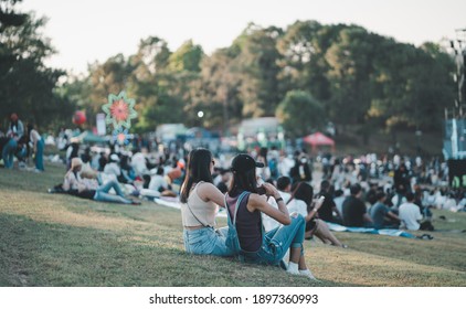 Girls friends watching concert in the park at open air, sitting in front of stage,spectators at background. 