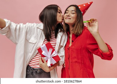 Girls In Good Mood Take Selfie On Pink Background. Cheerful Brunette Woman In White Oversized Shirt Kisses Her Friend And Holds Gift Box. Women Celebrating Birthday.