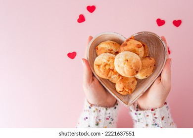 A Girl`s hand holding one Brazilian cheese bread in top view in a soft pink background with red little hearts and a heart shaped bowl