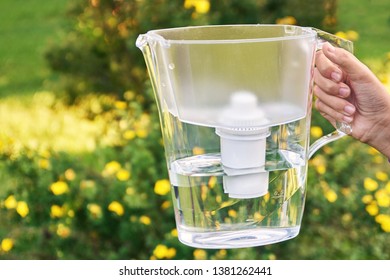 Girl's Hand Holding A Water Filtration Pitcher In The Sunny Summer Garden With Yellow Flowers On The Background In A Warm Day In Countryside