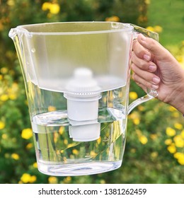 Girl's Hand Holding A Water Filtration Pitcher In The Sunny Summer Garden With Yellow Flowers On The Background In A Warm Day In Countryside