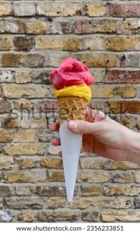 Girl's hand holds a beautiful ice cream. Part of the body. Brick wall on the background. Yellow and pink ice cream balls in a waffle cone. Hot summer day. Caucasian woman.