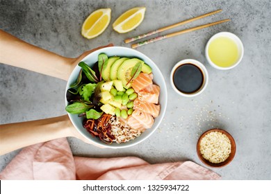 Girls' Hands Holding Salmon Poke Bowl Salmon Poke Bowl With Salad, Avocado, Edamame Beans, Pineapple, Sesame And Brown Rice Traditional Hawaiian Dish