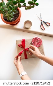 The Girl's Hands Tie A Red Ribbon Bow And Decorate A Gift For The Holiday. Gift Wrap. Kraft Paper Gift Box With Red Ribbon And Decor. The Process Of Wrapping A Gift On A White Table Next To Paper