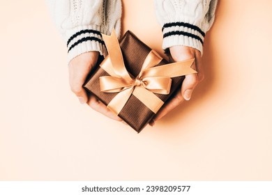 Girl's hands in white sweater holding brown gift box with golden bow on light peachy background. Christmas, Valentines day, Black friday sale concept. Top view. Peach Fuzz color of the year 2024