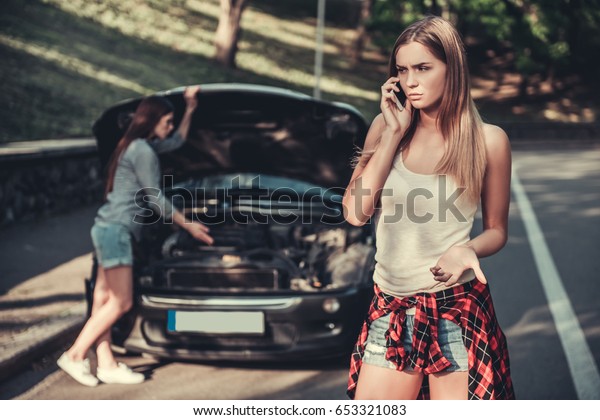 Girls having a\
problem with a car. One is talking on the mobile phone while\
another is studying motor\
hood