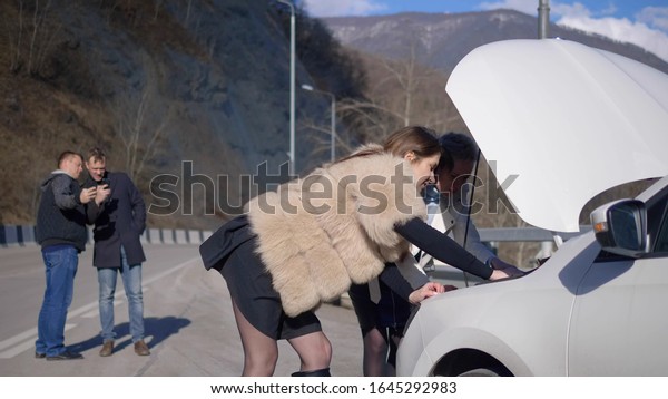 girls looks under the bumper of the car, men take\
it off on the phone.