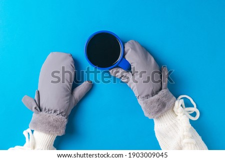 The girl's mittened hands hold a blue coffee mug on a blue background. Hot drink and mittens.