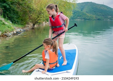 Girls Padding On Stand Up Paddle Boarding On Lake District. Children In Swim Life Vest Learning Paddleboarding On SUP Board. Family Active Leisure And Local Getaway With Kids Concept