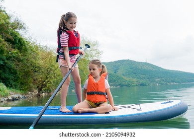 Girls Padding On Stand Up Paddle Boarding On Lake District. Children In Swim Life Vest Learning Paddleboarding On SUP Board. Family Active Leisure And Local Getaway With Kids Concept