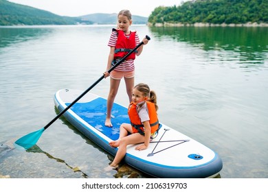 Girls Padding On Stand Up Paddle Boarding On Lake District. Children In Swim Life Vest Learning Paddleboarding On SUP Board. Family Active Leisure And Local Getaway With Kids Concept