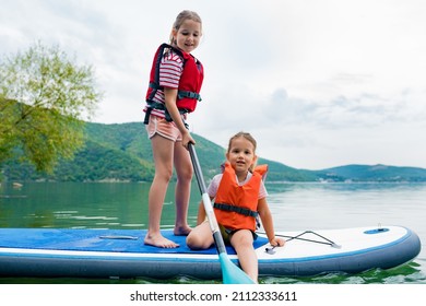 Girls Padding On Stand Up Paddle Boarding On Lake District. Children In Swim Life Vest Learning Paddleboarding On SUP Board. Family Active Leisure And Local Getaway With Kids Concept